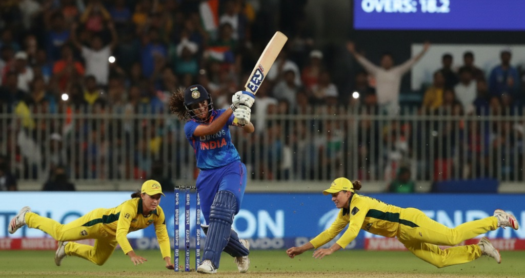 A high-energy moment from a women’s cricket match between India and Australia, showing Smriti Mandhana hitting a cover drive under stadium floodlights, with Australian fielders diving in the background, crowd cheering, and the scoreboard glowing — realistic photography style, professional sports photo.
